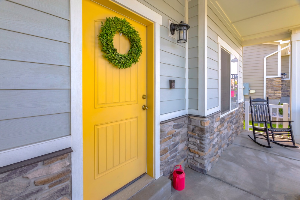 The porch of a light blue home with brick detail and a bright yellow front door accented by a green wreath